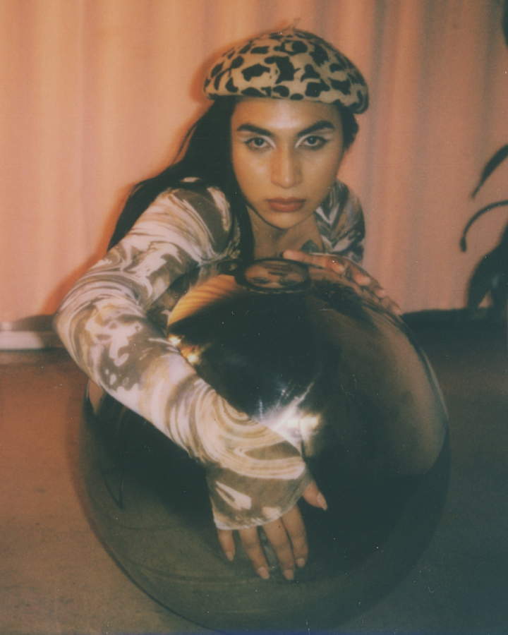 Model with cateye makeup on laying on the ground leaning against a metalic sphere looking at the camera